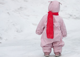 Small child in pink overalls with bunny ears lies and the bright scarf is walking on the snow in...