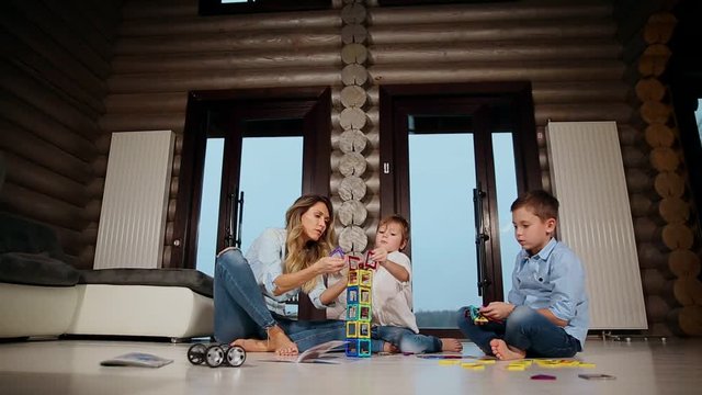 Happy mother with two children sitting on the floor of his country house spending time together collecting children's designer. Living room with panoramic Windows.
