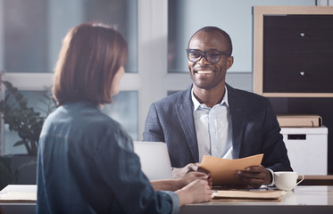 Glad to hear it. Portrait of optimistic young qualified african manager is looking at his colleague female with smile while sitting at table and having pleasant communication. Back view of woman - obrazy, fototapety, plakaty