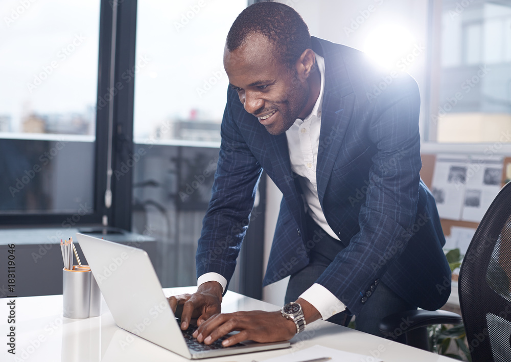 Poster writing project. cheerful successful adult african businessman in suit is typing on laptop with smil