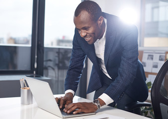 Writing project. Cheerful successful adult african businessman in suit is typing on laptop with smile. He is standing in office while leaning over table and working on gadget. Big window in background