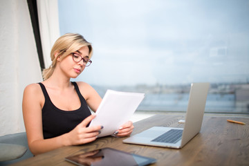 Businesswoman reading paper documents, preparing to the meeting with clients, sitting at work table with laptop computer and digital tablet. Female lawyer in glasses exploring summary or contract