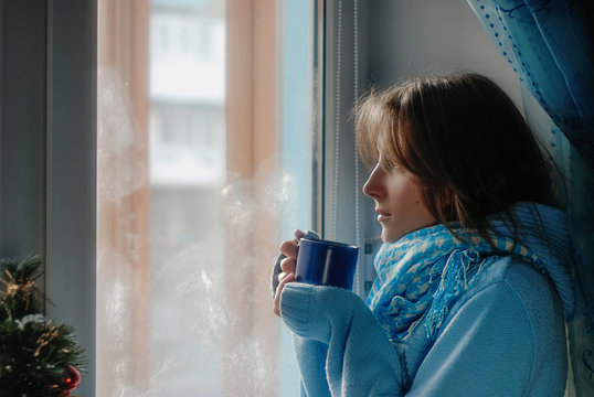 Sad upset lonly beautiful young woman with the cup in sweater dreaming near the frozen window at home