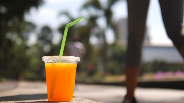 Cup of Tasty Fresh Orange Juice and Unrecognized Running Man on Background in Morning City Park. 4K, Slow Motion. Thailand.