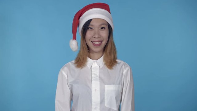 young asian woman posing wearing santa claus hat on blue background in studio. attractive millennial girl wearing white casual shirt looking at the camera laughing