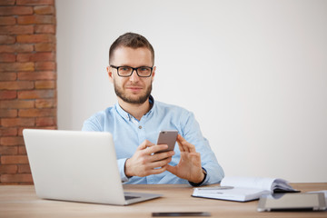 Close up of mature unshaven male manager in glasses and shirt sitting in office, working on laptop computer, chatting on smartphone, looking in camera with satisfied expression.