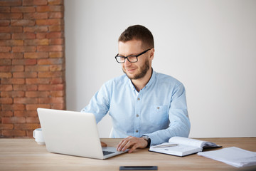 Portrait of cheerful handsome unshaven male company manager in glasses and casual clothes sitting...