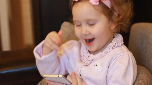 A little cute girl sitting in sofa in a living room, looking cartoon and playing the game. Funny child looks at the phone screen and plays downloaded application on a smart phone