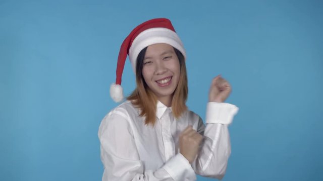 young asian woman posing wearing santa claus hat dancing on blue background in studio. attractive millennial girl wearing white casual shirt looking at the camera laughing