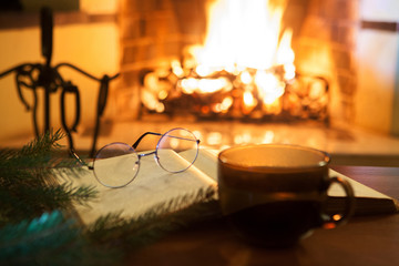 An open ancient book, a cup with a drink in front of the fireplace. Evening fairy tales. Fantasy. Christmas.