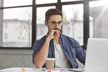 View of concentrated talented skilled male focused on screen of laptop computer, reads necessary information for creating budget report, drinks hot beverage. Attractive male freelancer keyboards text
