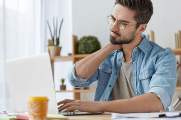 Overworked male student works all day on course paper, has pain in neck, has sedentary life, tired of sitting on one place, keyboards on laptop computer, drinks coffee. People, work, tiredness concept