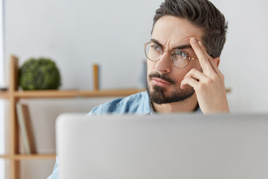 Horizontal shot of bearded stylish male copywriter thinks about content of new article, sits in front of opened laptop, looks thoughtfully aside, has serious expression. People and work concept