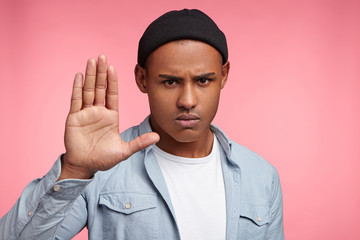 Portrait of serious sunburnt young hipster guy shows stop sign, asks not do something or refuses, rejects action, has strict look, isolated over pink background. Stylish man wears hat says no gestures