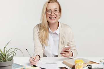 Indoor shot of smiling businesswoman has pleased happy expression, writes notes or fills documents, holds smart phone, searches examples of making contracts or statute as wants to open new company