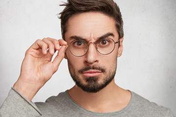 Close up portrait of confident bearded man looks indignant through round spectacles, raises eyebrows, listens to news or interlocutor, poses against white studio background. Facial expressions