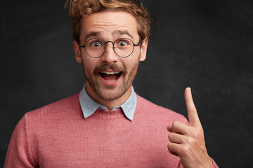 Excited bearded male nerd looks with amazed expression, gets brilliant idea, raises fore finger,...