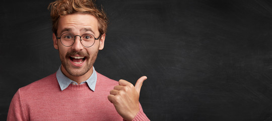 Cheerful male nerd in spectacles stands against chalkboard, points with thumb on copy space for your advertising content. Horizontal shot of glad college professor shows something at blackboard - obrazy, fototapety, plakaty