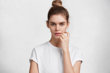 Serious content beautiful young female with hair knot, wears casual white t shirt, enjoys domestic atmosphere, spends weekends at home, poses against white studio background. Beauty concept.