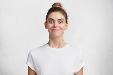 Fototapeta premium Cheerful female model with happy expression and charming appearance, has dark hair tired in knot, glad to meet handsome guy, isolated over white background. Emotional young beautiful woman indoor