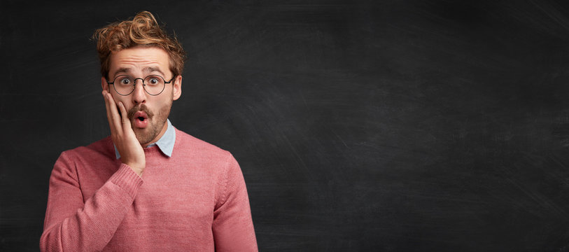 Amazed Young Male Post Graduate Student Looks With Shocked Expression, Realized That Tomorow Is Presentation Of Dissertation Or Defend Of Thesis, Poses Against Chalk Black Board With Copy Space