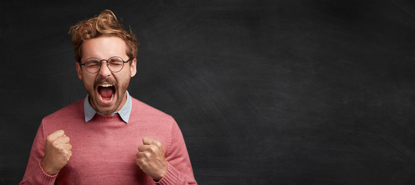 Emotional stylish guy, clenches fists rejoices his great success, screams with enjoyment and happiness isolated on black chalk background. People, reaction, emotions and body language concept