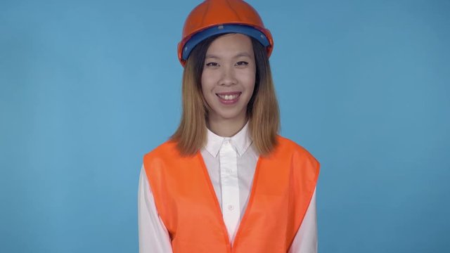 young asian woman posing on blue background in studio. attractive millennial girl wearing white casual shirt and orange hard hat and vest looking at the camera with cheerful happy smile