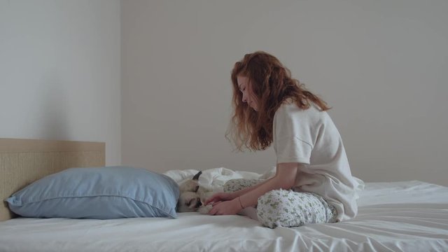 young redhead girl in pajamas in the morning playing with the kitten in the bedroom on a white bed, holiday home with Pets