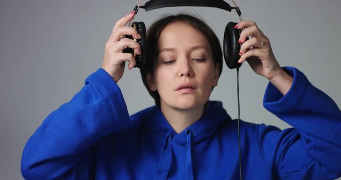 Calm woman in a casual hoodie with short dark hair puts on large overhead headphones and starts smiling and dancing to music