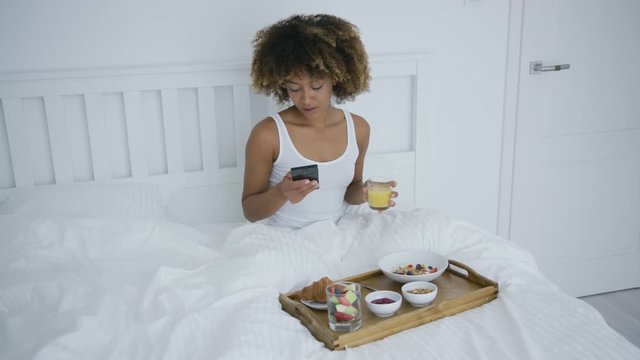 Pretty ethnic model in white top sitting in bed having tray with served breakfast near and drinking juice while watching smartphone in moning.