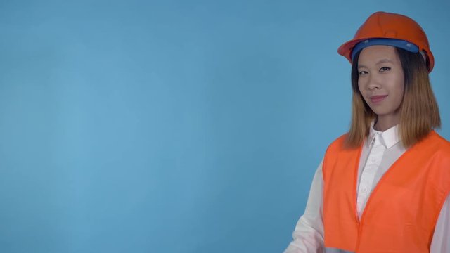 young asian woman posing pointing on copy space on blue background in studio . attractive millennial girl wearing white casual shirt and orange hard hat and vest looking at the camera with cheerful
