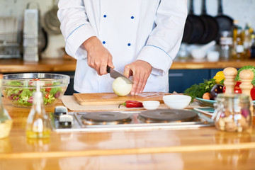 Unrecognizable male chef wearing uniform standing at table of restaurant kitchen and cutting onion while preparing vegetarian salad