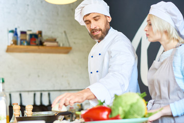 Handsome bearded chef wearing uniform explaining pretty woman how to prepare vegetable soup...