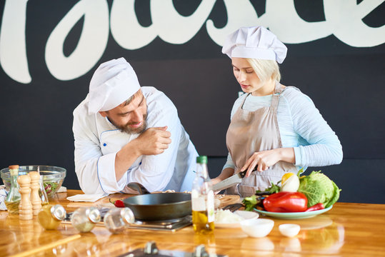 Concentrated young woman wearing apron cutting vegetables on wooden board while smiling middle-aged chef keeping eye on her, interior of restaurant kitchen on background