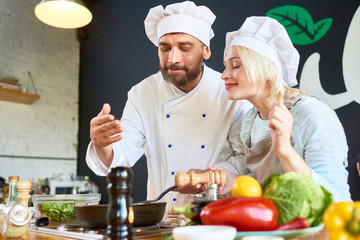 Experienced bearded chef and his attractive young assistant enjoying smells coming from frying pan while preparing delicious dish at restaurant kitchen