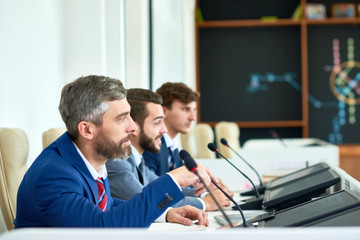 Profile view of confident entrepreneurs wearing elegant suits sitting in row at table and answering...