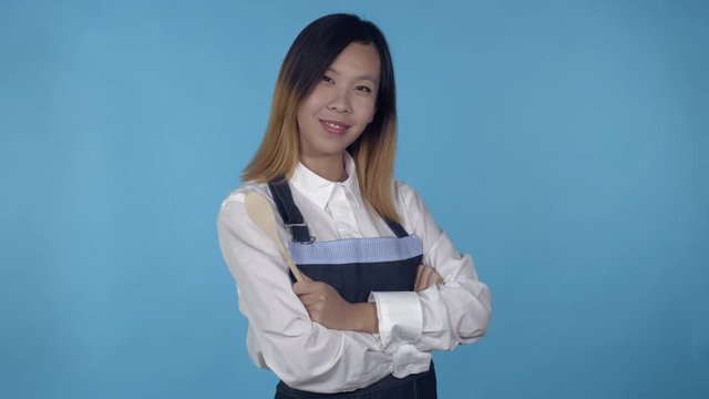young asian woman posing with spoon wearing like cook on blue background in studio. attractive millennial girl wearing white casual shirt looking at the camera smiling.