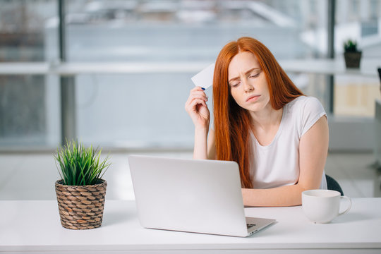 Online Payment Shopping Concept. Thinking Business Woman Sitting At Table With Laptop, Holding Empty Credit Card, Looking Away In Thoughts