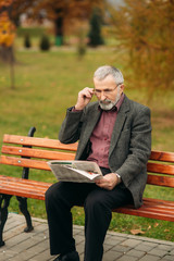 Fototapeta premium A nice grandfather with a beautiful beard in a gray jacket sits on a bench in the park and reads a newspaper