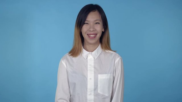 young asian happy woman posing showing positive emotions on blue background in studio. attractive millennial girl wearing white casual shirt looking at the camera smiling