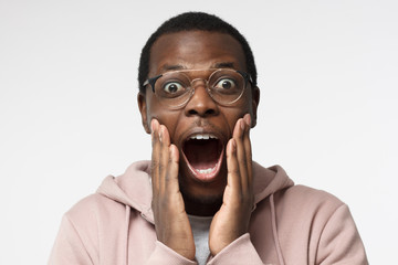 Closeup shot of young handsome African guy in casual clothes pictured isolated on white background, wearing round spectacles, looking extremely surprised with round eyes and mouth, positively shocked