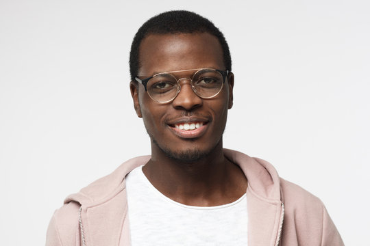 Indoor portrait of young handsome African male pictured isolated on white background wearing white T-shirt, pink hoodie and round eyeglasses showing relaxed smile and looking confident and satisfied