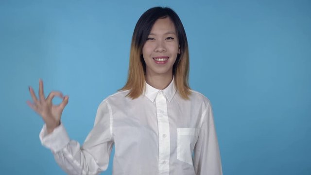 young asian woman posing showing hand gesture ok on blue background in studio. attractive millennial girl wearing white casual shirt looking at the camera smiling