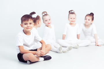 Little ballerinas and kid ballerun doing exercises and sitting on floor in white ballet class.