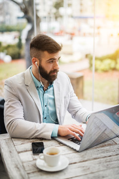 Close up portrait of young handsome attractive bearded businessman sitting at the table in a suit and typing on a laptop while looking far away and having earphones for a coffee break outside.
