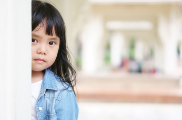 Asian children cute or kid girl peek with sneaks behind the pole and wear jeans for fashion with make face scowl and sad or angry at pavilion garden on holiday with space