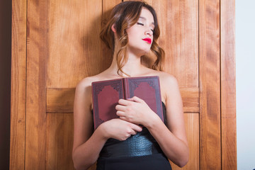     Beautiful young girl in negligee sitting and holding book in room, eyes closed 