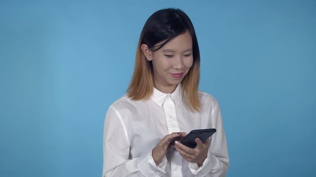 young asian woman using smartphone for app or social media on blue background in studio. attractive millennial girl wearing white casual shirt looking at the camera smiling