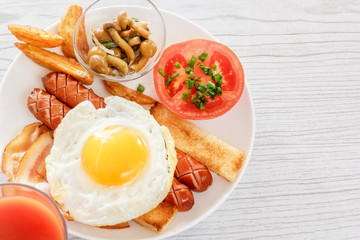 Full English breakfast with smoked sausages, scrambled eggs, bacon, tomatoes, mushrooms, toast and beans. A glass of fresh juice. Breakfast on a white plate on a light background. Space for text. 