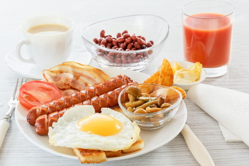 Full English breakfast with smoked sausages, fried egg, bacon, tomato, toast and beans. Tea with milk. A glass of fresh juice. Cutlery. Breakfast on a white plate on a light background. 
